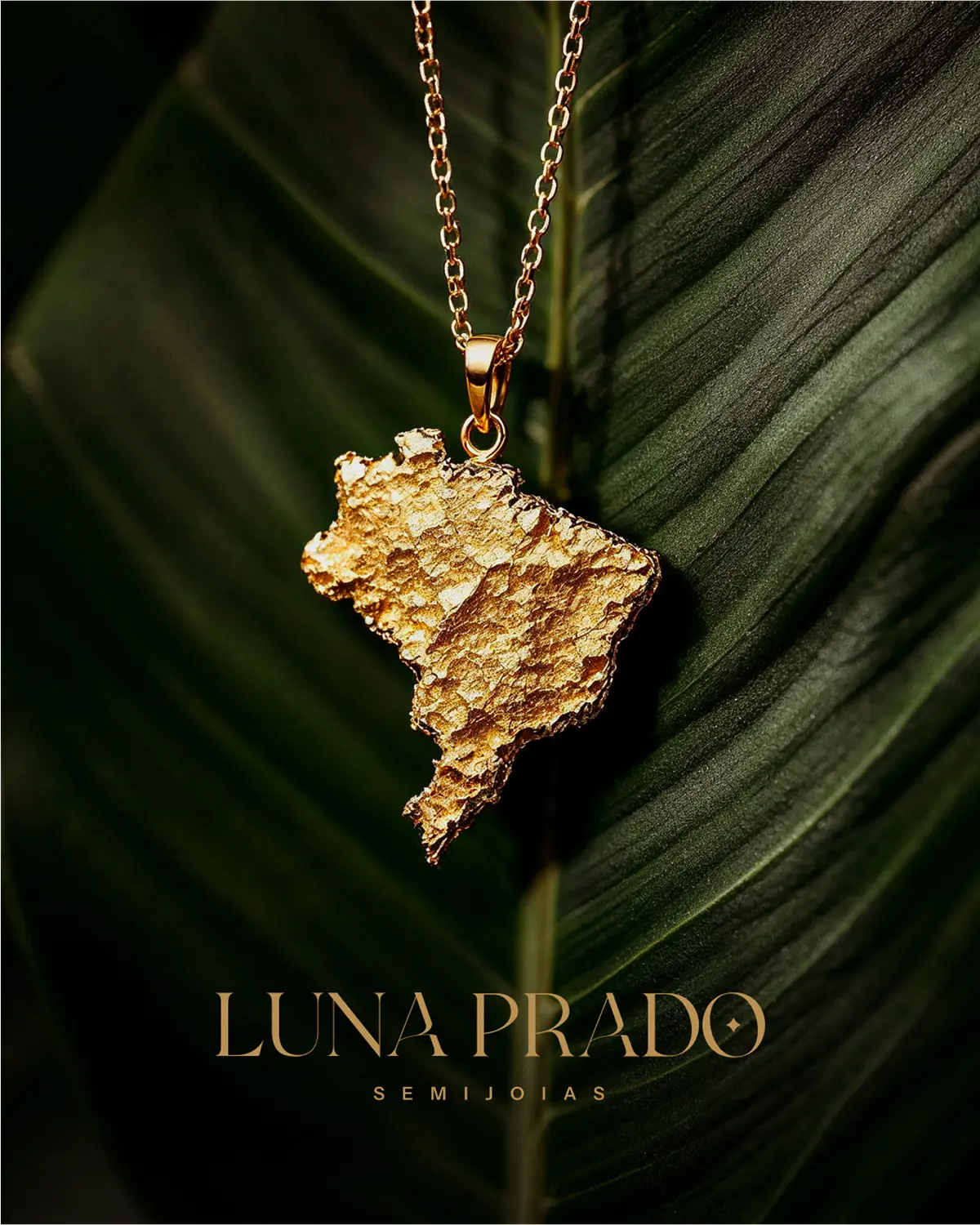 Colar dourado com pingente do mapa do Brasil sobre folha verde, com o logo de Luna Prado.